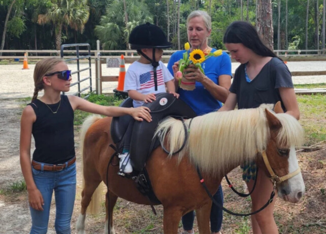 Four people and a pony. Two girls, a woman, and a young child, are gathered around the pony in an outdoor setting with trees and fencing. The young child is sitting on the pony wearing a riding helmet, while a woman holds a bouquet of flowers and hel