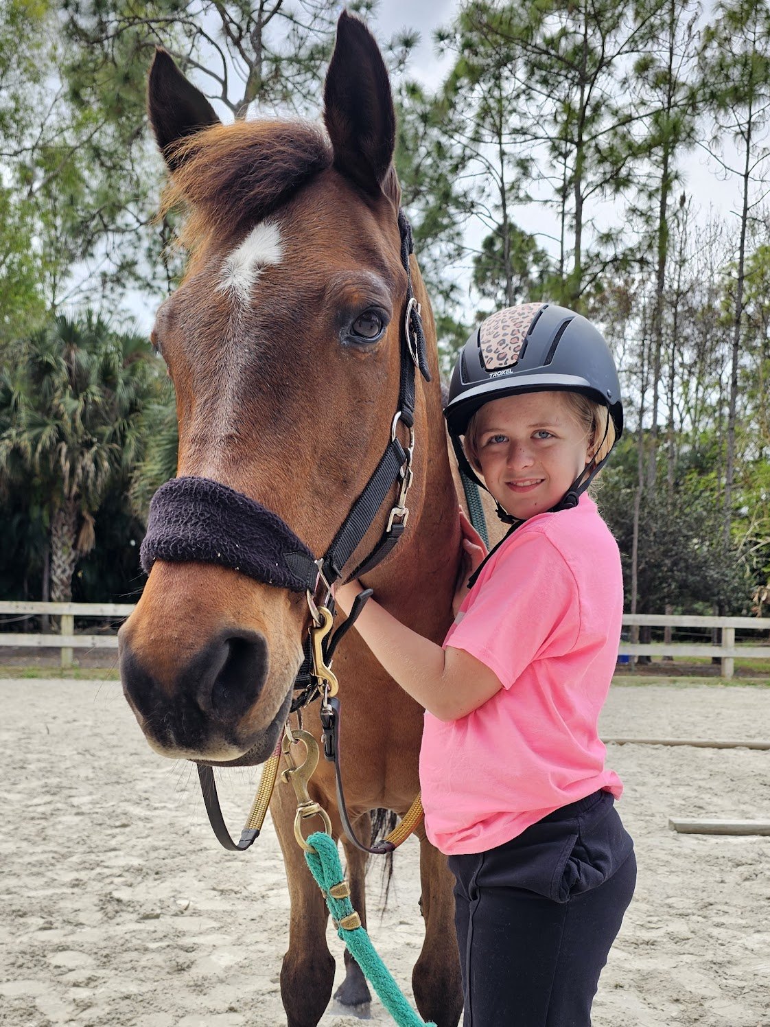 A young girl wearing a pink shirt and a protective helmet is hugging a brown horse with a white marking on its face at an outdoor riding arena.