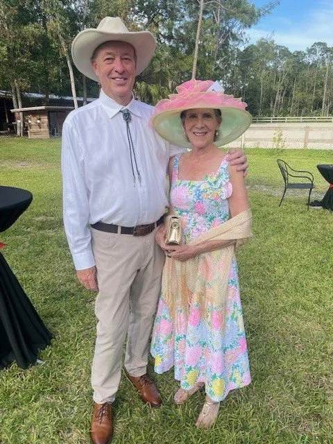 A smiling elderly man and woman dressed in vintage-style clothing and large wide-brimmed hats, standing outdoors in a grassy area with trees. The man wears a white shirt, tan pants, and a cowboy hat. The woman wears a floral dress, a pink hat, and ho