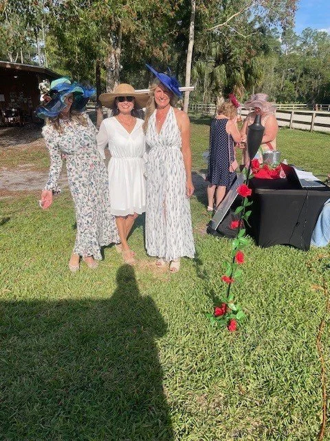 Three women in floral and white dresses, wearing large hats, standing outdoors on grass near a table and other people at a garden or outdoor event.