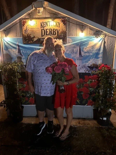 A man and woman standing together, holding a bouquet of pink flowers, at a Kentucky Derby event with a decorated backdrop featuring roses and Derby themed signs.