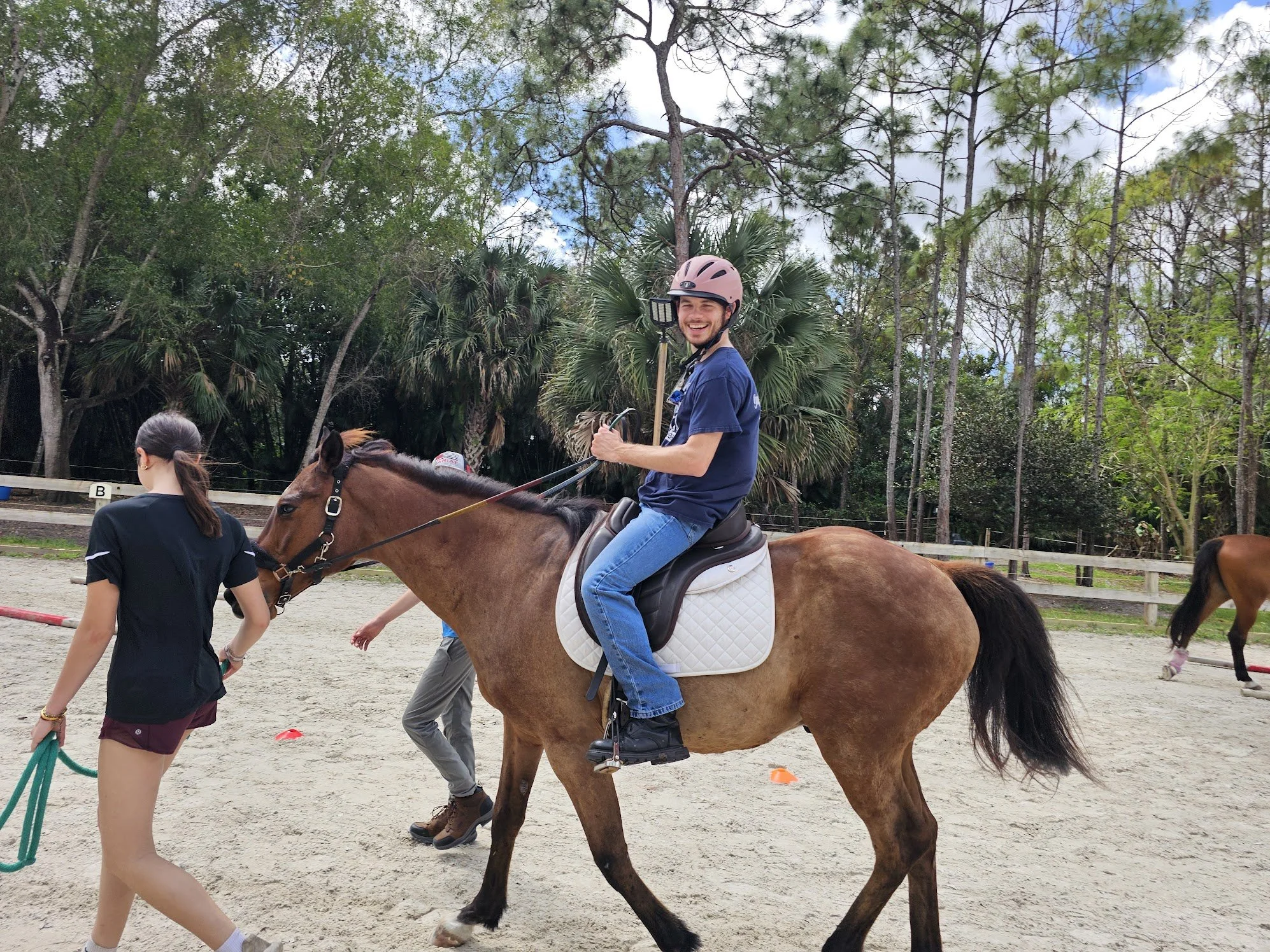 A man in a pink helmet riding a brown horse at an outdoor equestrian facility, with a smiling woman leading the horse.