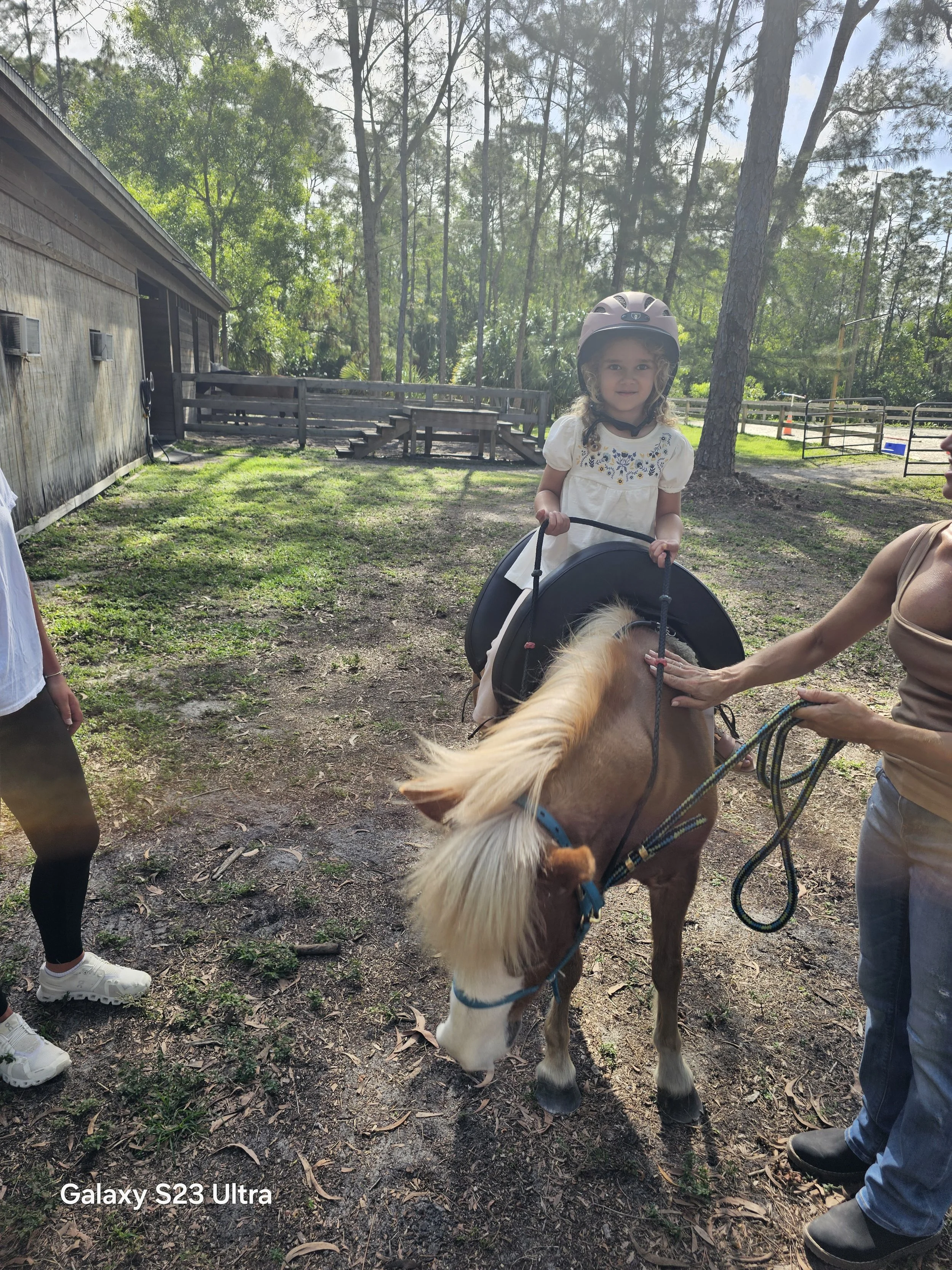 A young girl sitting on a small horse and wearing a helmet, being held by a woman on the right with a lead rope, in an outdoor setting with trees and a wooden building in the background.