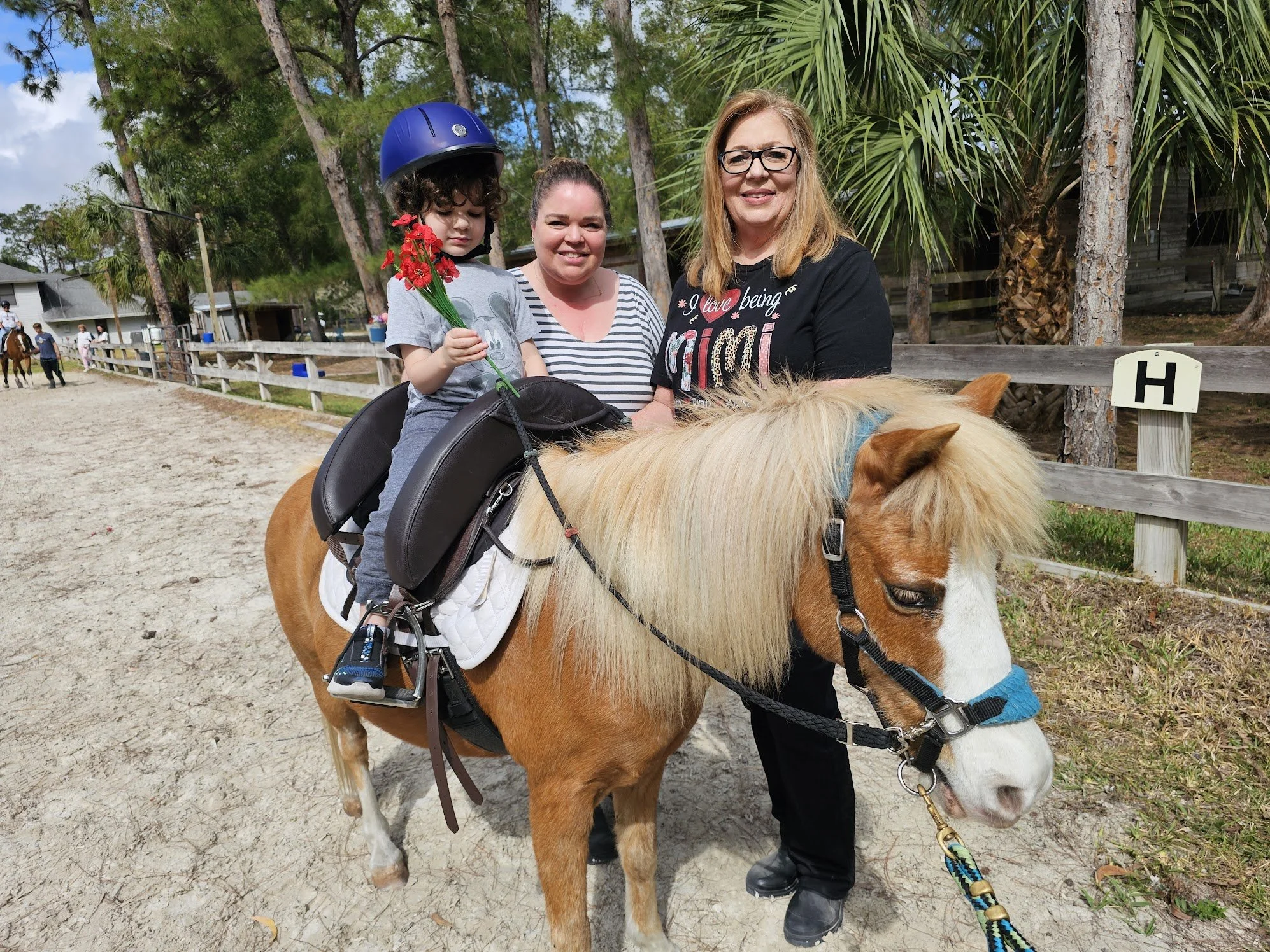 A woman, a child with a helmet, and another woman standing next to a pony in an outdoor setting with trees and a wooden fence.