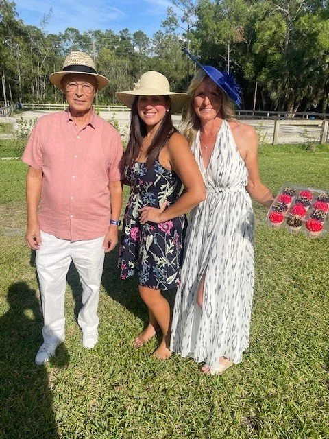Three people standing outdoors on grass, all wearing hats and smiling. The man on the left wears a light pink shirt, white pants, and a straw hat. The woman in the middle wears a floral dress and a beige hat. The woman on the right wears a striped dr