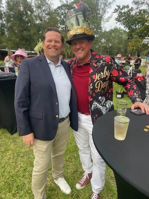 Two men standing outside at a gathering, one wearing a suit and the other in a festive hat and colorful jacket, with drinks on a table nearby.