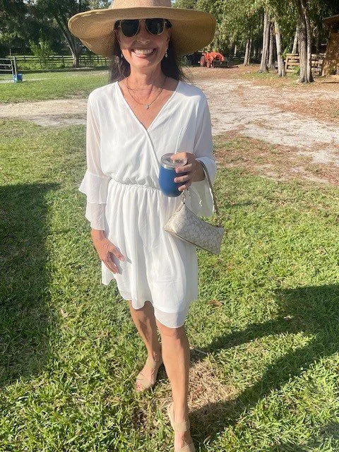 A woman stands outdoors on a grassy area, wearing a white dress, large brimmed straw hat, and sunglasses, holding a beverage and a purse.