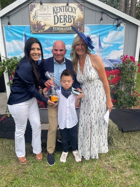 Four people standing outdoors at a Kentucky Derby fundraiser event, with a banner above them that says 'Kentucky Derby Fundraiser'. They are smiling, and three of them are holding glasses of beer. There are some flowering plants behind them.