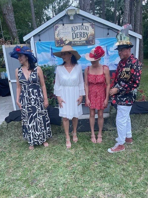 Four people dressed in fancy, colorful outfits and large, elaborate hats standing in front of a sign that reads 'Kentucky Derby Fundraiser' outside with trees in the background.