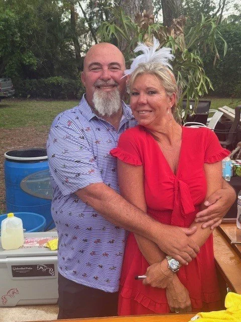 A man and woman embracing outdoors, smiling, with greenery in the background. The woman is wearing a red dress and white feathered headpiece, and the man is wearing a patterned shirt.