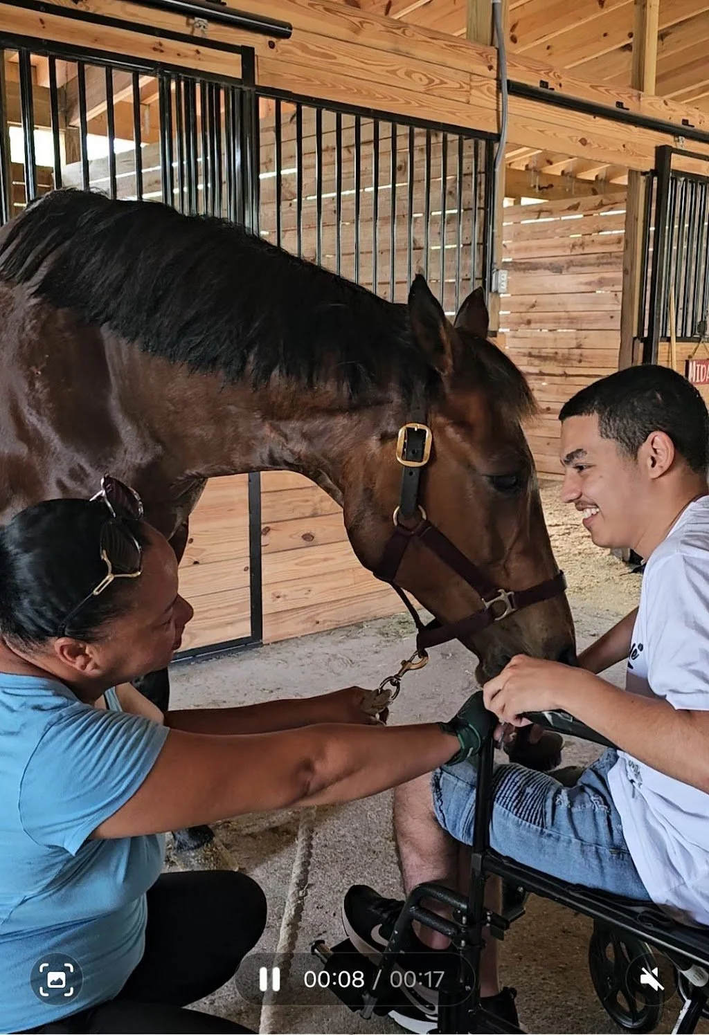 A man in a wheelchair and a woman in a blue shirt are smiling and holding a horse's head in a barn with wooden walls and stalls in the background.