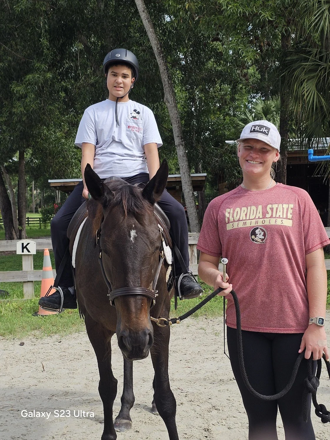 A young boy is riding a horse with a woman standing next to it, holding the reins. The boy is wearing a helmet and a light grey t-shirt, while the woman is wearing a cap and a red t-shirt with 'Florida State Seminoles' written on it. The setting appe