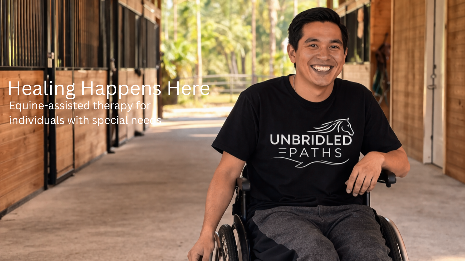 A smiling man in a wheelchair, wearing a black T-shirt with the logo "Unbridled Paths," sitting inside a stable or barn with wooden walls and stalls.