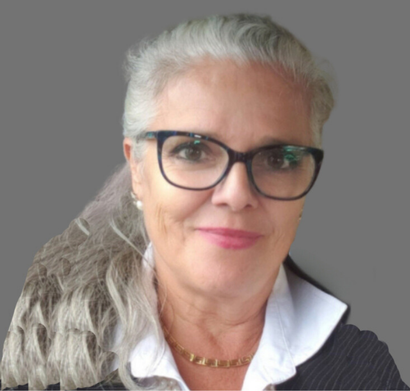 A woman with long gray hair, glasses, and wearing a white collared shirt and black blazer, smiles at the camera against a gray background.