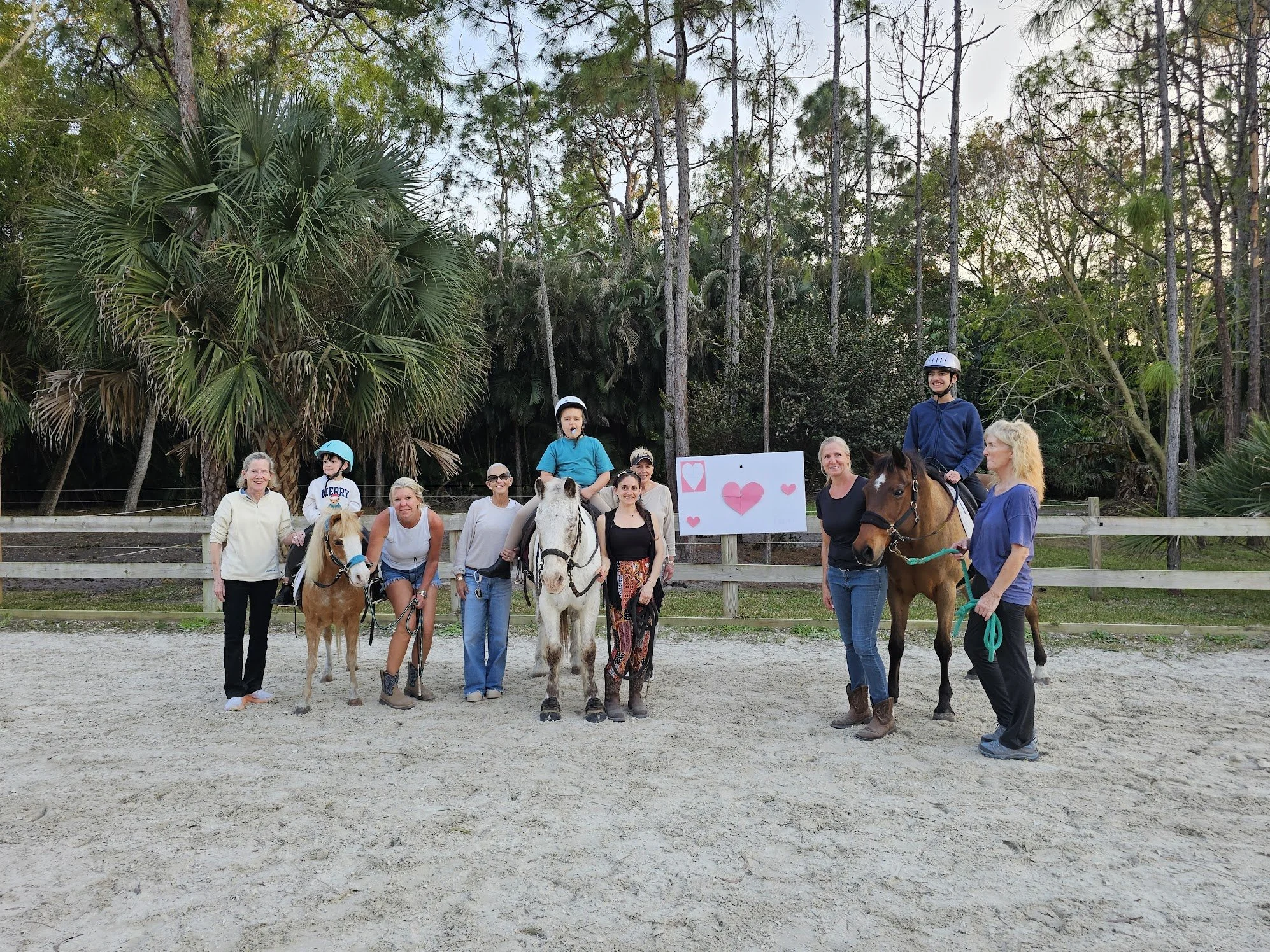 Group of people, including children, adults, and seniors, standing and riding horses in an outdoor equestrian area with trees in the background, decorated with a pink heart-themed sign.
