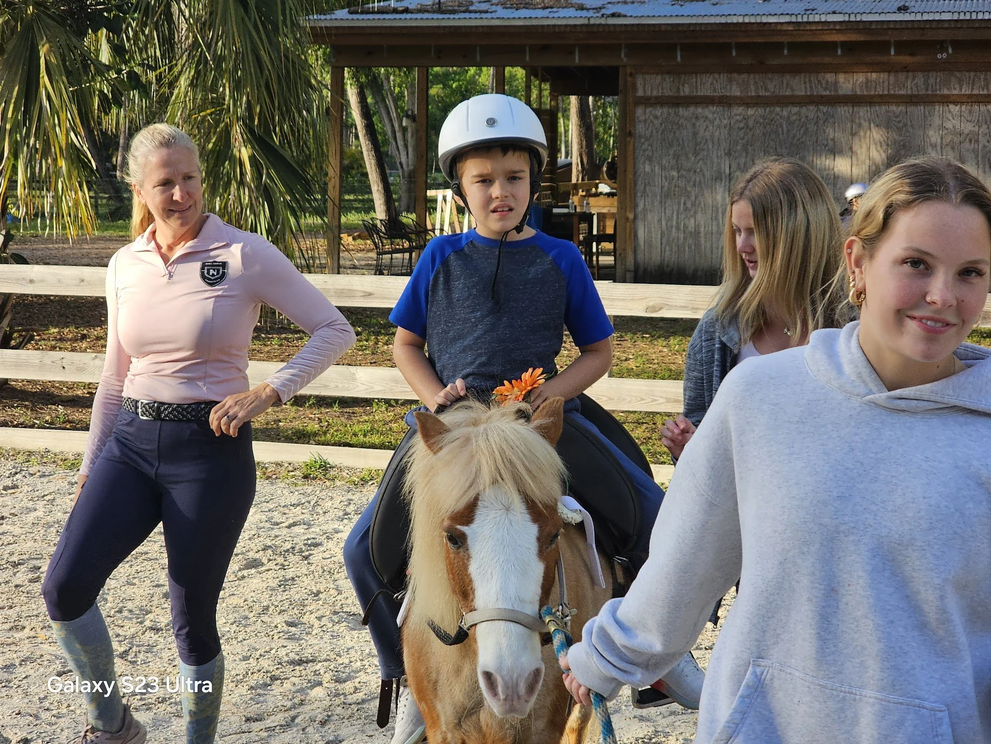 A young boy wearing a helmet is sitting on a small horse or pony, while a woman stands nearby with her hand on her hip, and a girl in a gray hoodie is holding the horse's lead, with two other women in the background. The scene appears to be at a hors