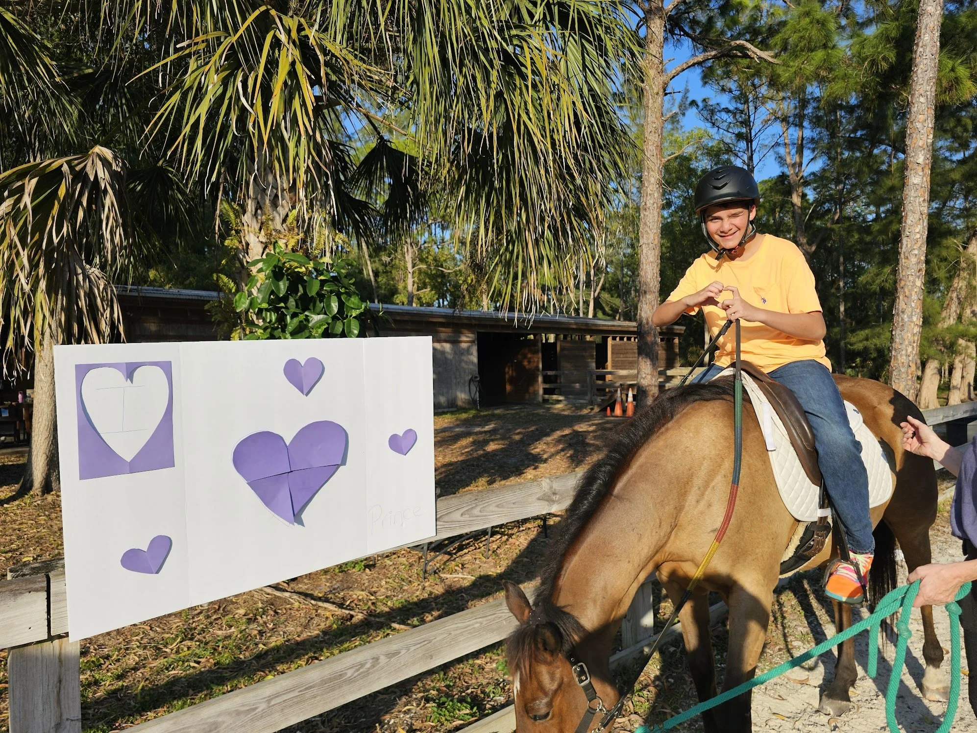 A young girl wearing a black riding helmet and yellow shirt is riding a brown horse along a wooden fence. She is smiling and holding the reins. Near them, there is a sign with purple paper hearts attached to the fence, with some hearts featuring the 