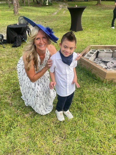 A woman and a boy are smiling outdoors near a fire pit at a gathering or event. The woman is kneeling beside the boy, who is standing. The woman is wearing a large blue hat and a white dress with black pattern. The boy is dressed in a white shirt, da