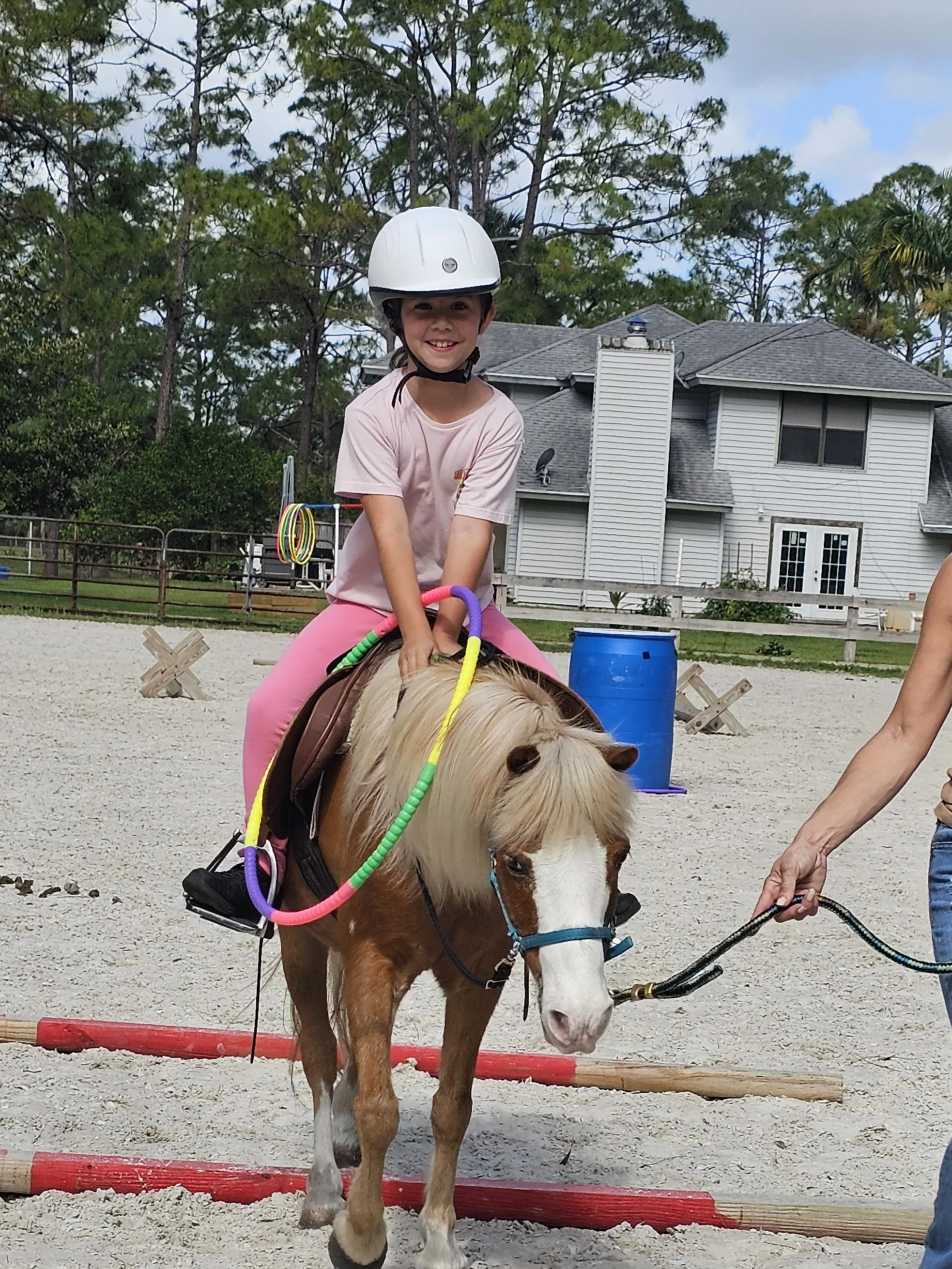 A young girl wearing a white safety helmet riding a small horse with a blonde mane. The girl is smiling and holding the horse's reins, while an adult holds an additional lead rope. The scene takes place in an outdoor riding arena with a residential h