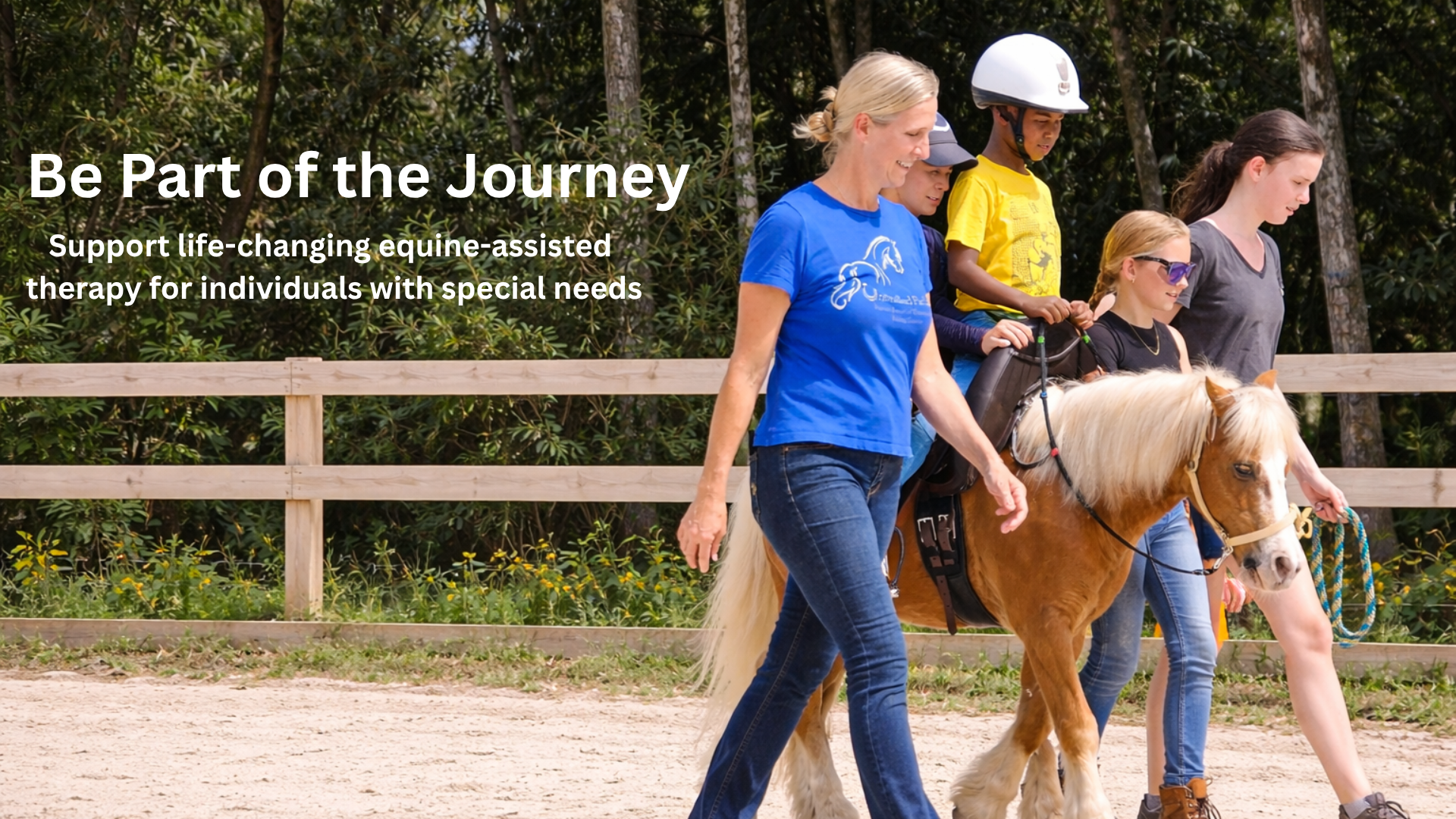 A woman leading a small horse with three young girls riding a horse on a dirt path with a wooden fence and green trees in the background.
