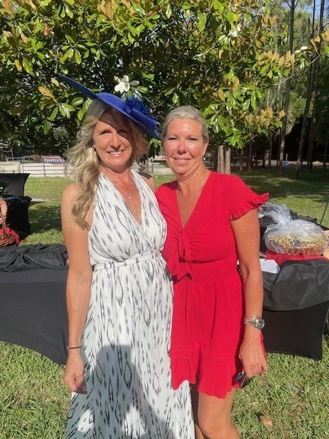 Two women smiling outdoors under a tree; one in a white patterned dress and a blue hat, the other in a red dress.