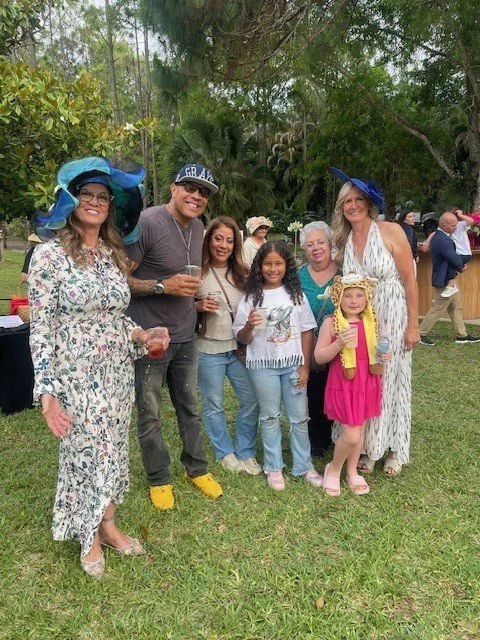 Group of seven people, including adults and children, dressed in summer clothes and hats, standing outdoors in a grassy area with trees, smiling and posing for a photo.