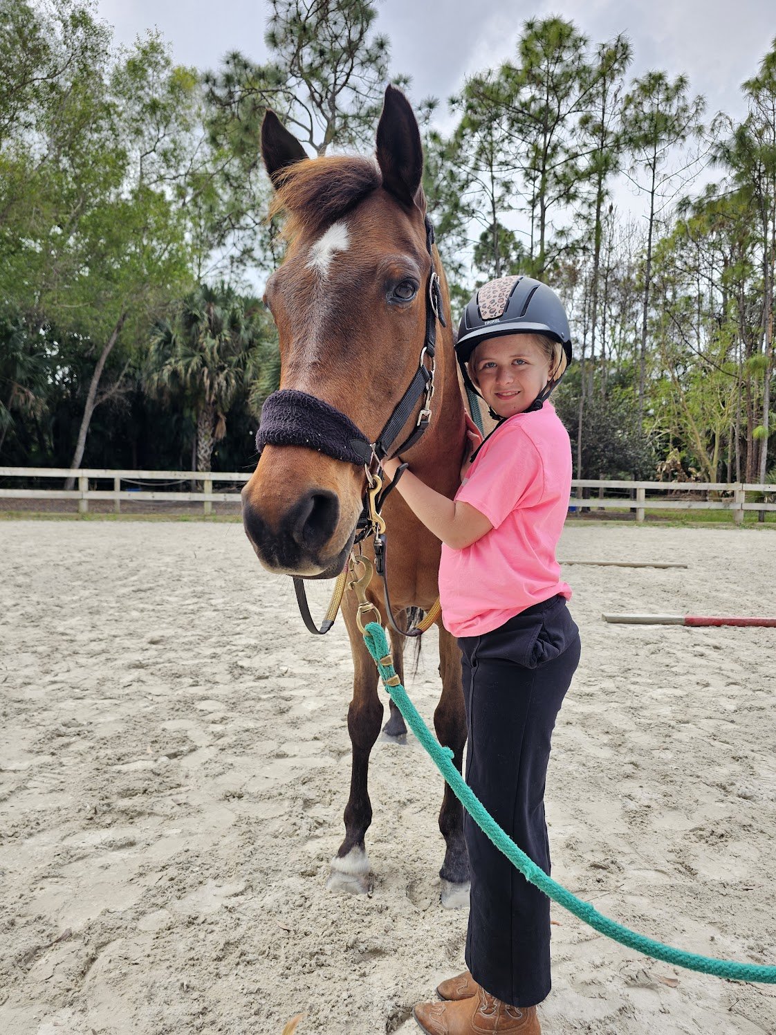 A young girl wearing a pink shirt, black pants, and a riding helmet standing next to a brown horse with a white marking on its face. The girl is holding the horse's bridle and smiling. They are in an outdoor riding arena with sand footing and a woode