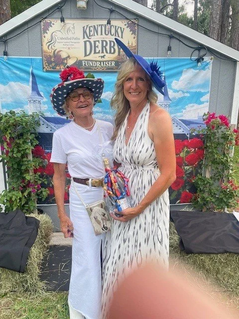 Two women dressed in festive attire standing in front of a Kentucky Derby fundraiser banner, with floral decorations around them.