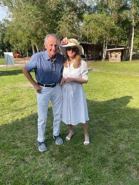An older man and woman standing on grass in a park during daytime, smiling at the camera. The man wears a short-sleeved blue shirt, white pants, and gray shoes. The woman wears a white dress, wide-brimmed hat with pink flowers, and white heels. Trees