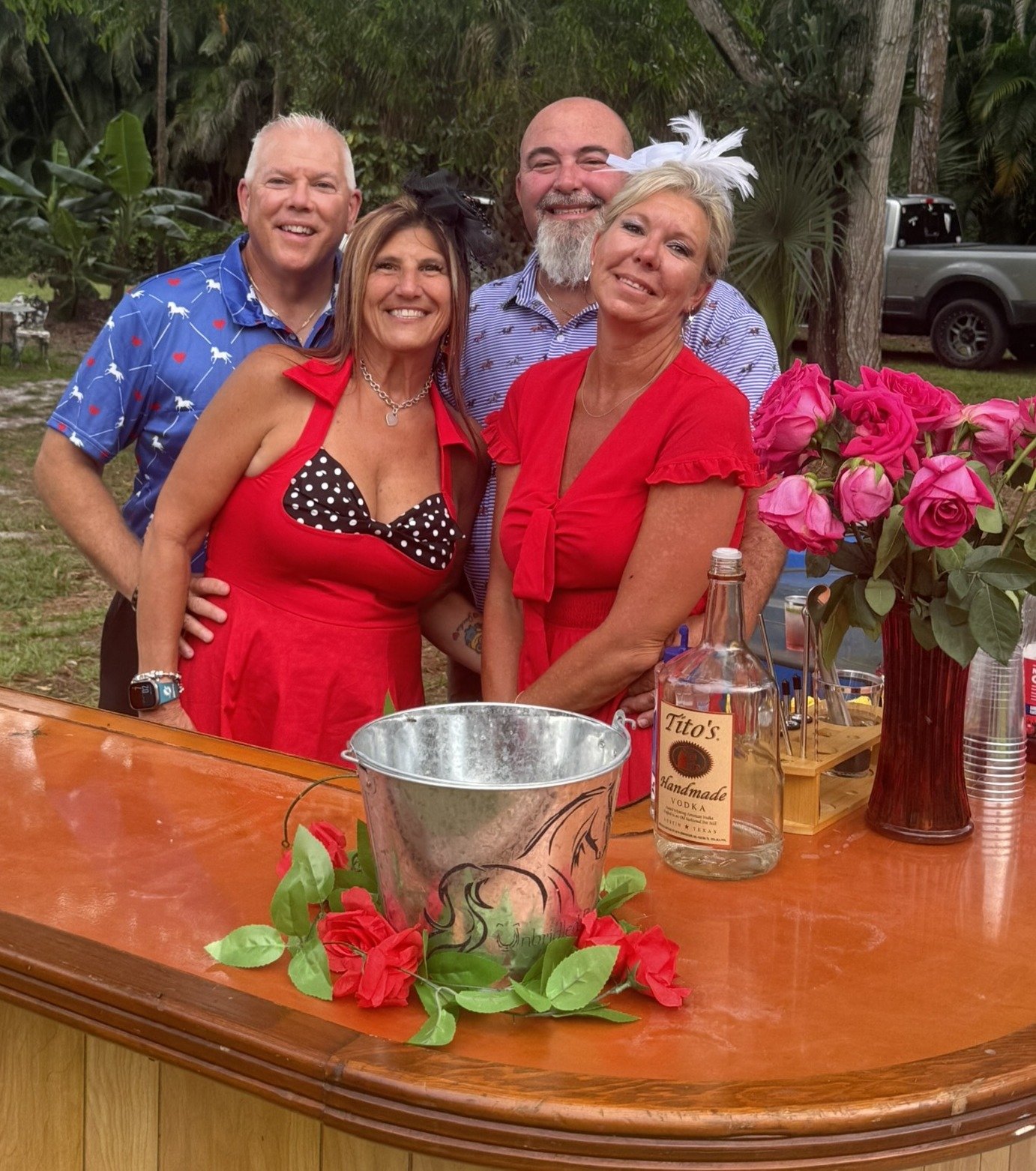 A group of five smiling people standing behind a wooden bar table decorated with pink roses and green leaves. The table has a metal bucket, a bottle of Tito's Handmade Vodka, and a vase with pink roses. The background features trees and a parked truc