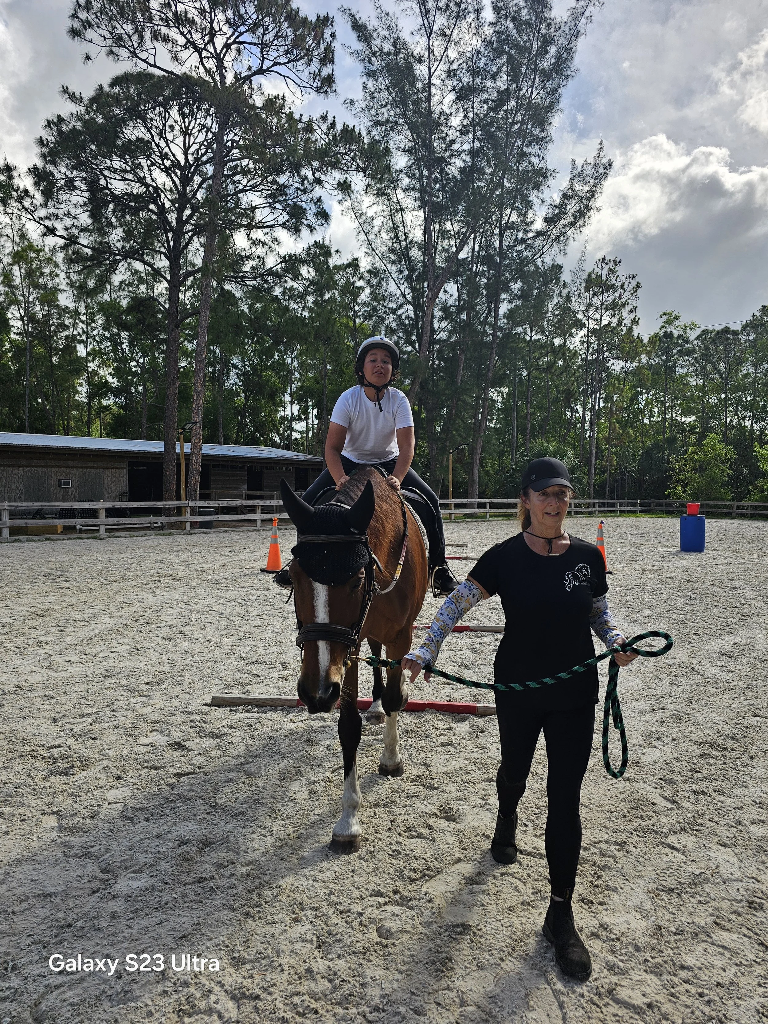 A young girl riding a horse with a woman leading the horse in an outdoor riding arena surrounded by trees.