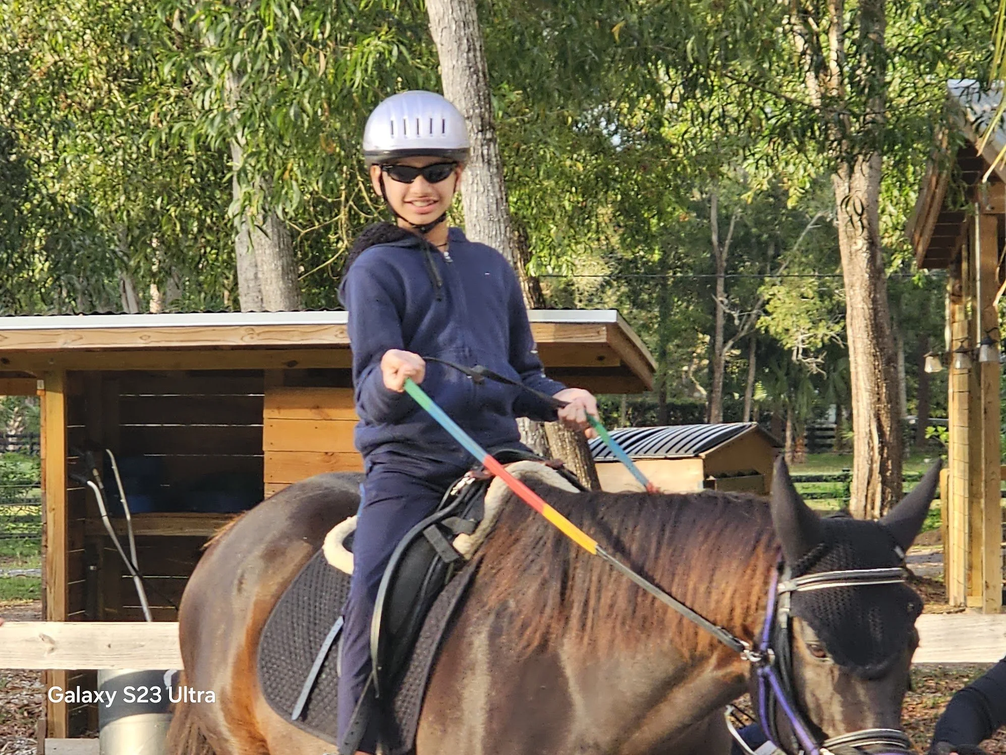 A person riding a horse, wearing a riding helmet and sunglasses, smiling, in a wooded outdoor setting.