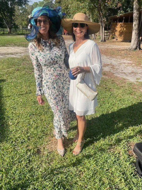Two women standing outdoors on grass, dressed in elegant summer outfits with large hats and sunglasses, smiling at the camera.