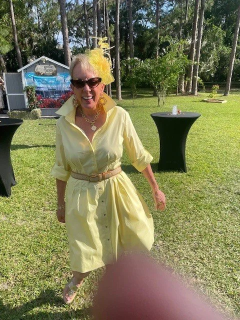 A smiling woman dressed in a yellow dress with rolled-up sleeves, sunglasses, a yellow feathered fascinator, and a necklace, standing outdoors on a grassy lawn.