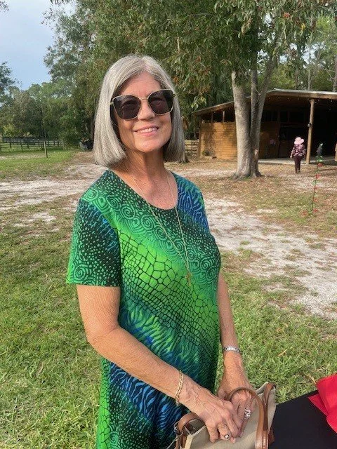 A smiling older woman with gray hair, wearing black sunglasses and a vibrant, patterned green and blue dress, standing outdoors in a grassy park area with trees and a small wooden building in the background.