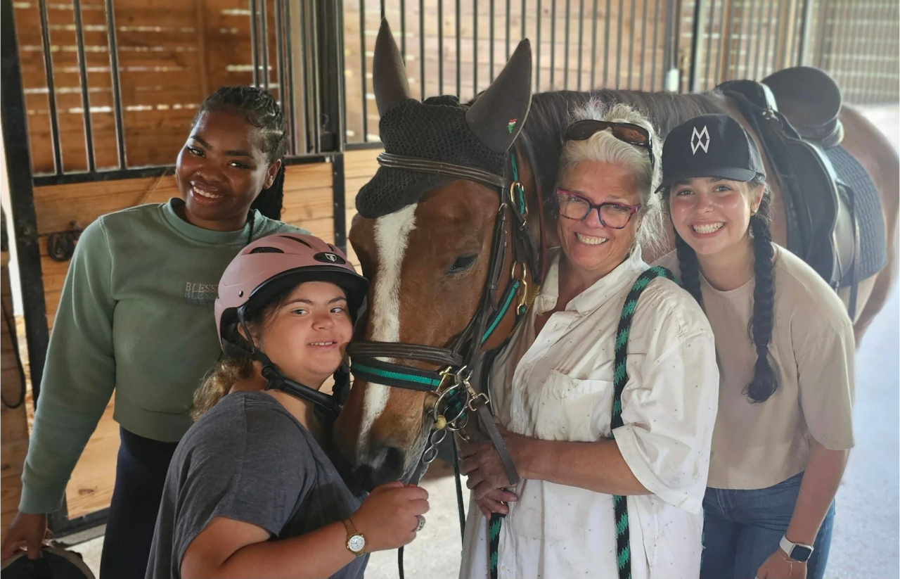 Four women and a horse inside a wooden stable, with two women wearing riding helmets and two women with glasses and braids, all smiling and close to the horse.