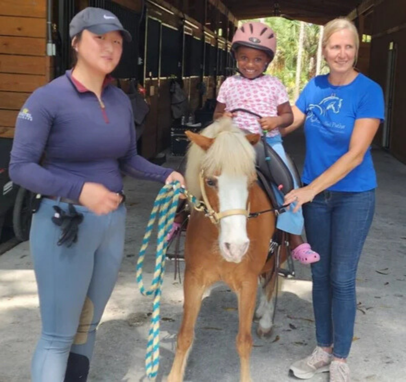 A young girl wearing a pink helmet sitting on a small horse, flanked by two women, one on each side, in a stable or barn setting.