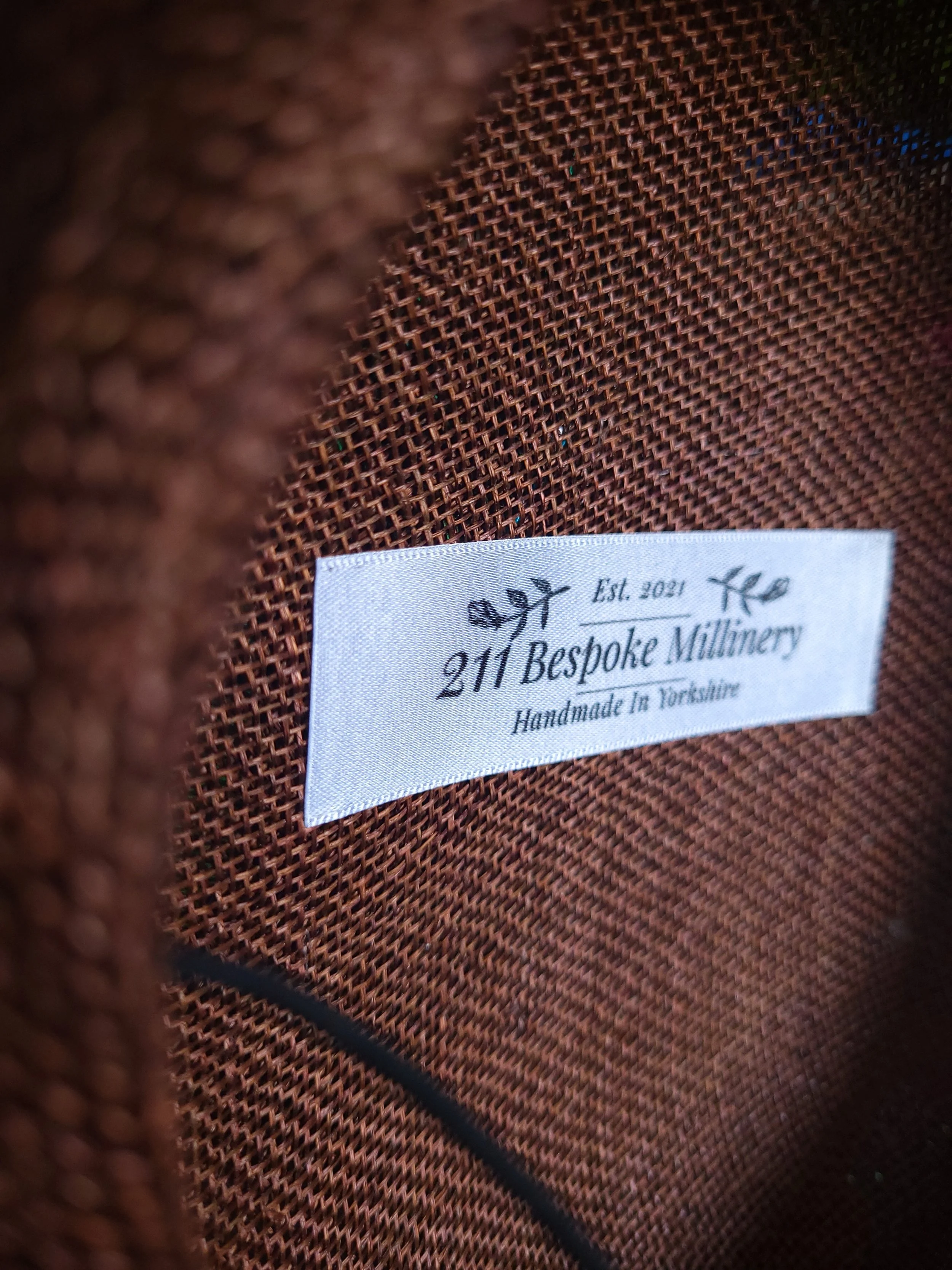 Close-up of a white fabric label inside a brown textured item, displaying the text '& Est. 2021 211 Bespoke Millinery Handmade in Torquay' with decorative floral symbols.