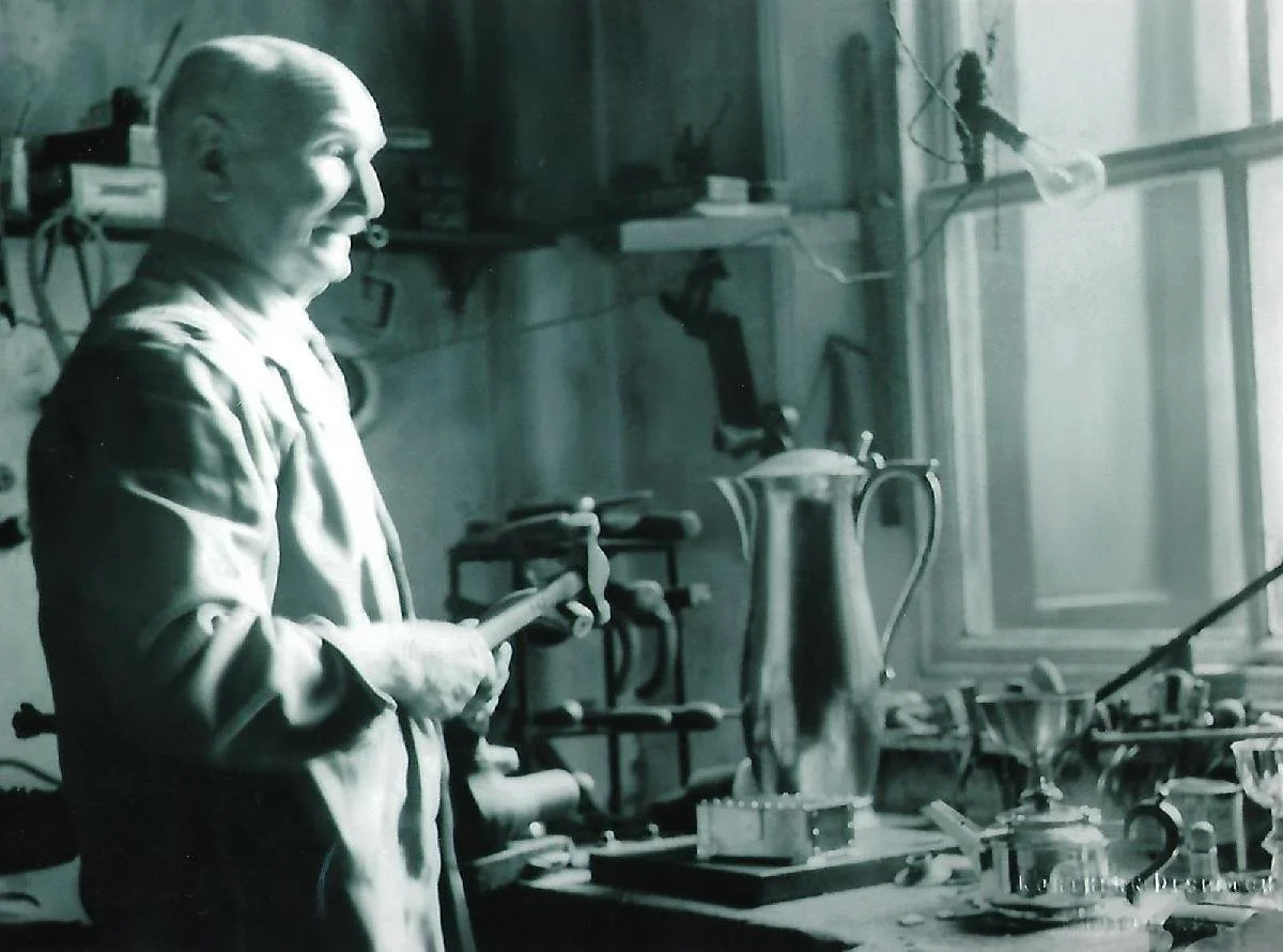 An elderly man in a workshop, holding a hammer, standing near a workbench with a large metal jug, various tools, and objects, with a window in the background.