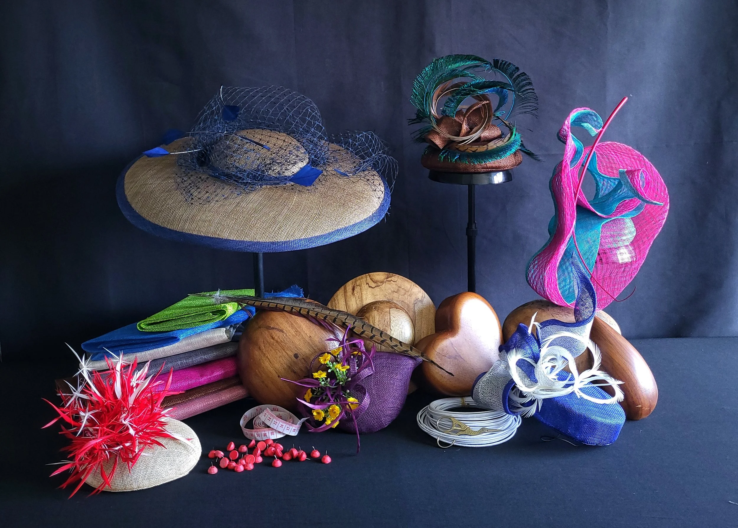 Collection of various hats and headpieces displayed on black surface with a dark background. The hats include a wide-brimmed straw hat with blue netting, a smaller fascinator with green and brown feathers, and a pink and blue mesh hat. Other items in