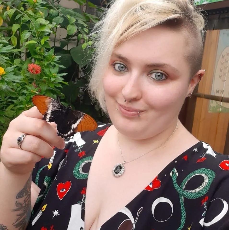 A woman with light skin, blue eyes, and short blonde hair with an undercut, holding an orange and black butterfly, standing outdoors with greenery and wooden structures in the background.