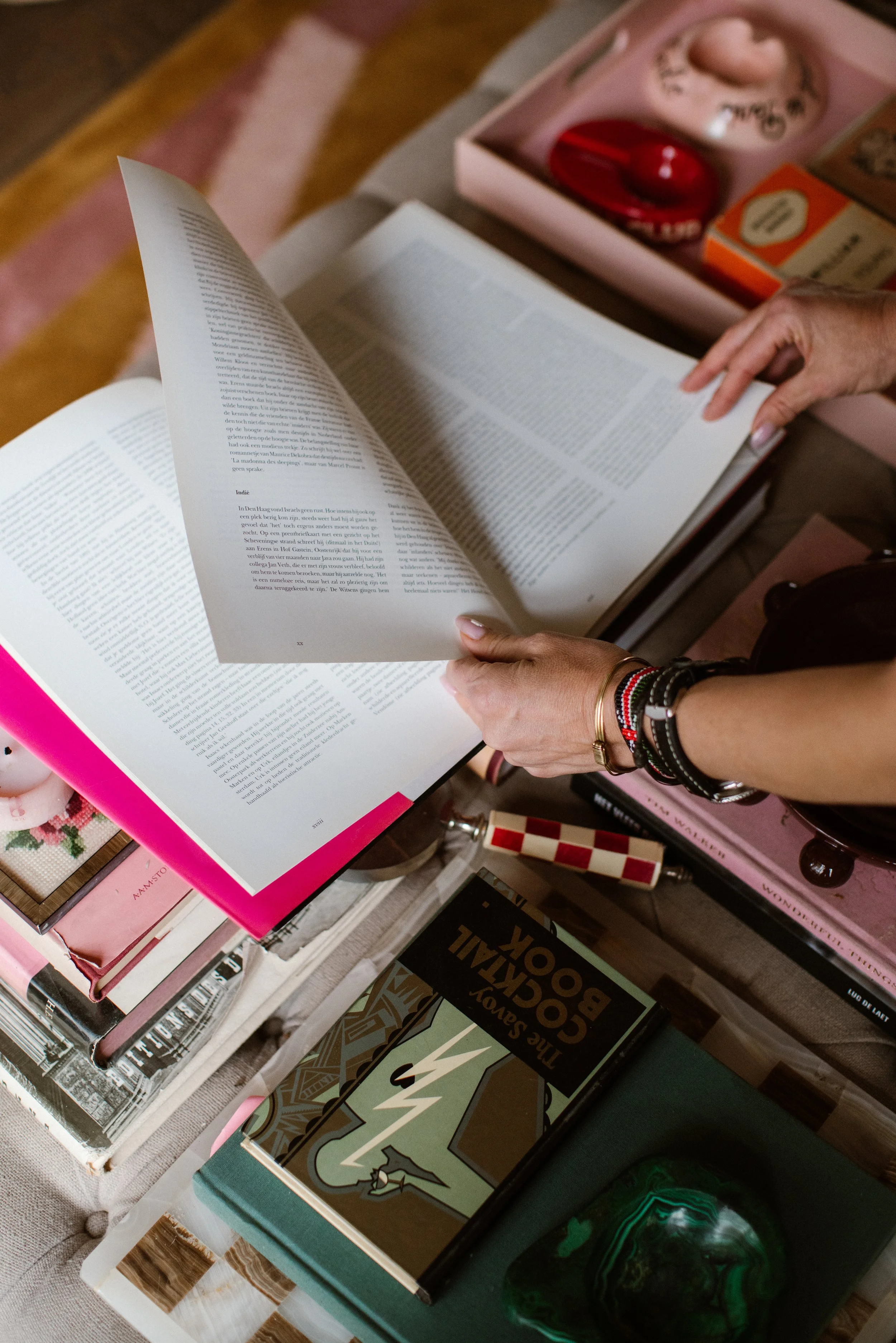 A person flipping through a paperback book with carefully manicured nails. Various books are stacked on a table, including titles such as "The Social Book" and "The Art of Laquer."