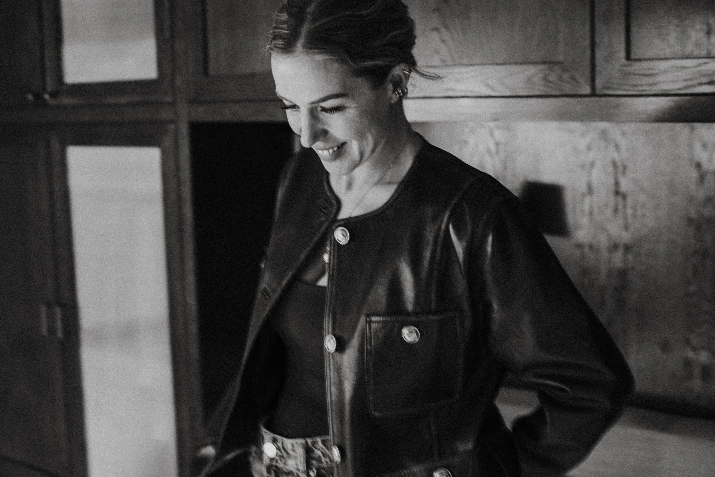 A woman with dark hair and earrings smiling, wearing a leather jacket and a top, standing against a wooden wall, captured in black and white.