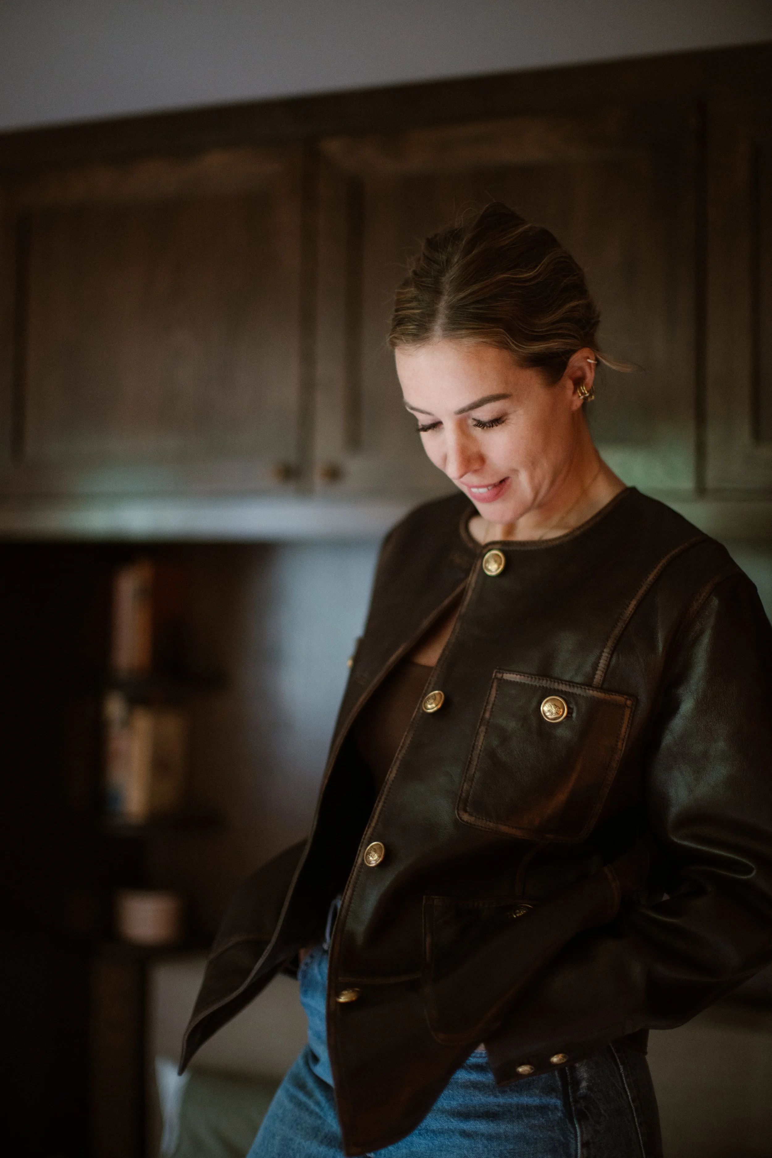 A woman with brown hair styled in waves, wearing a black leather jacket with gold buttons and blue jeans, smiling looking downward in a home kitchen.