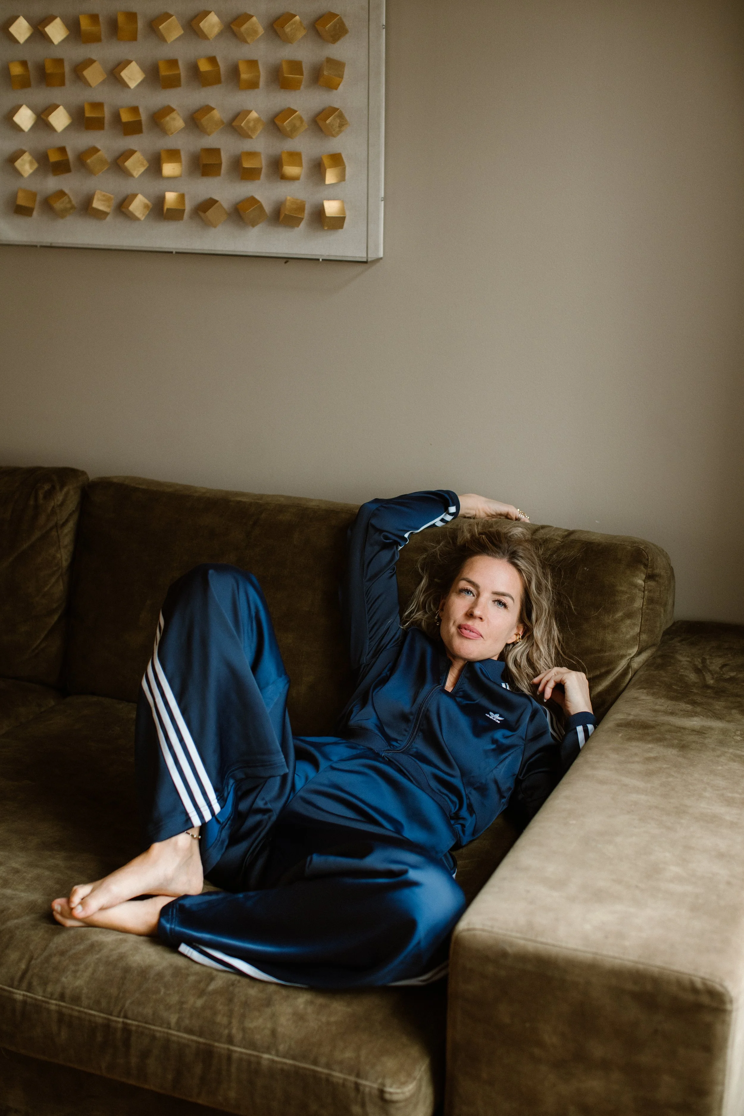A woman with blonde curly hair lying on a brown couch in a relaxed pose, wearing a navy blue tracksuit with white stripes, in a room with beige walls and a wall-mounted art piece consisting of small gold cubes.