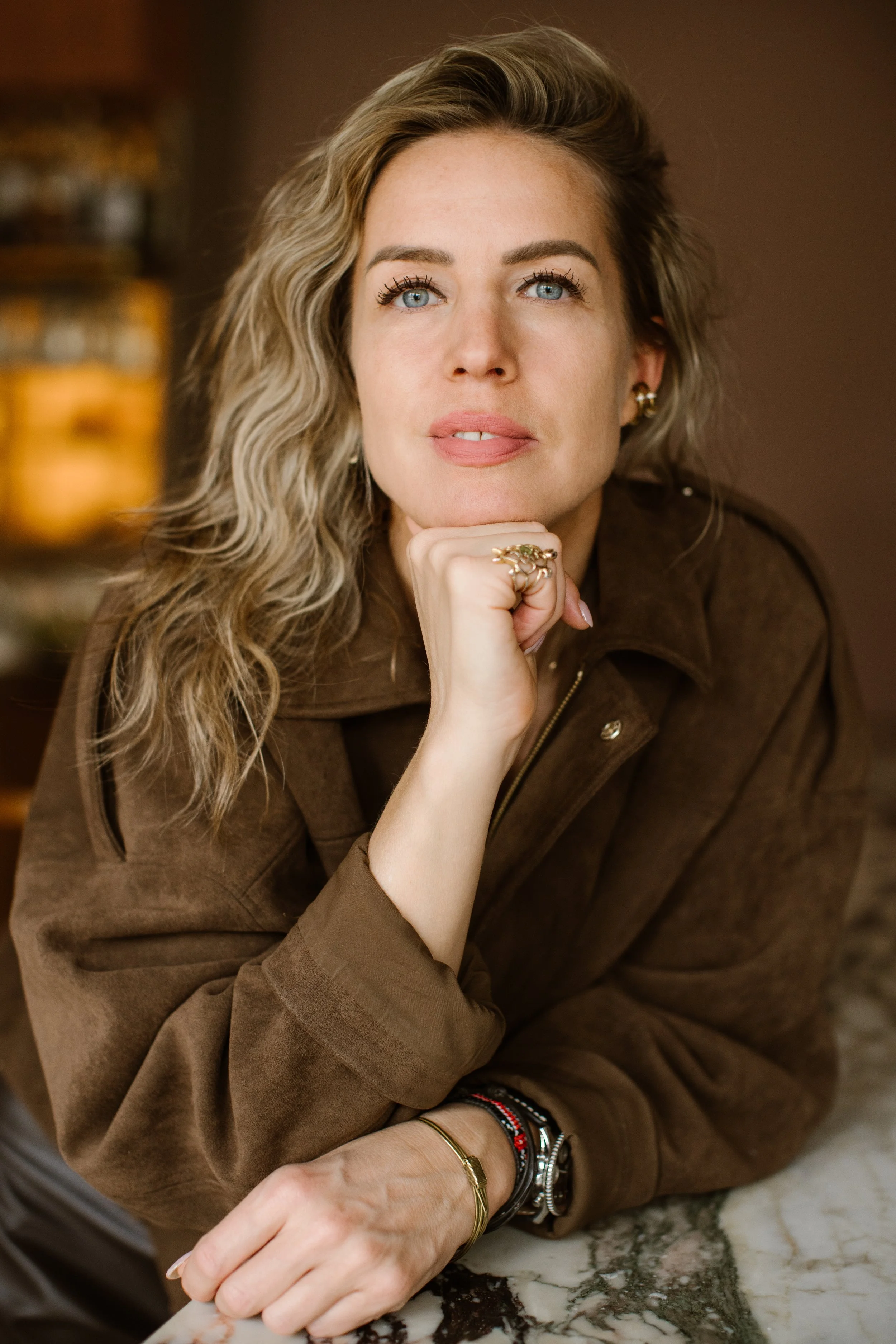 A woman with curly blonde hair and blue eyes resting her chin on her hand, wearing a brown jacket and various jewelry, leaning on a marble surface.