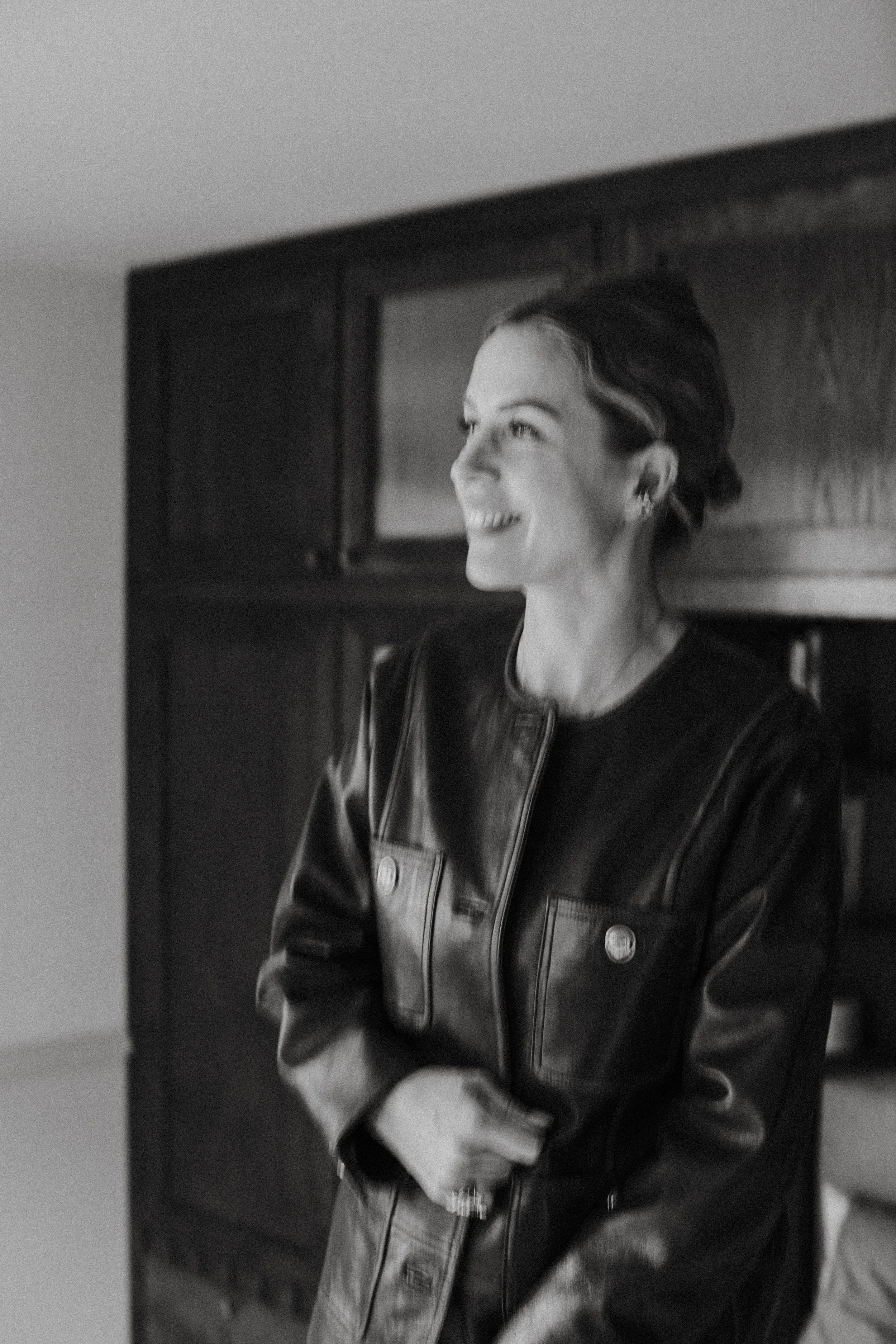 Black and white photo of a smiling woman with short wavy hair wearing a leather jacket, standing in front of a dark bookshelf.
