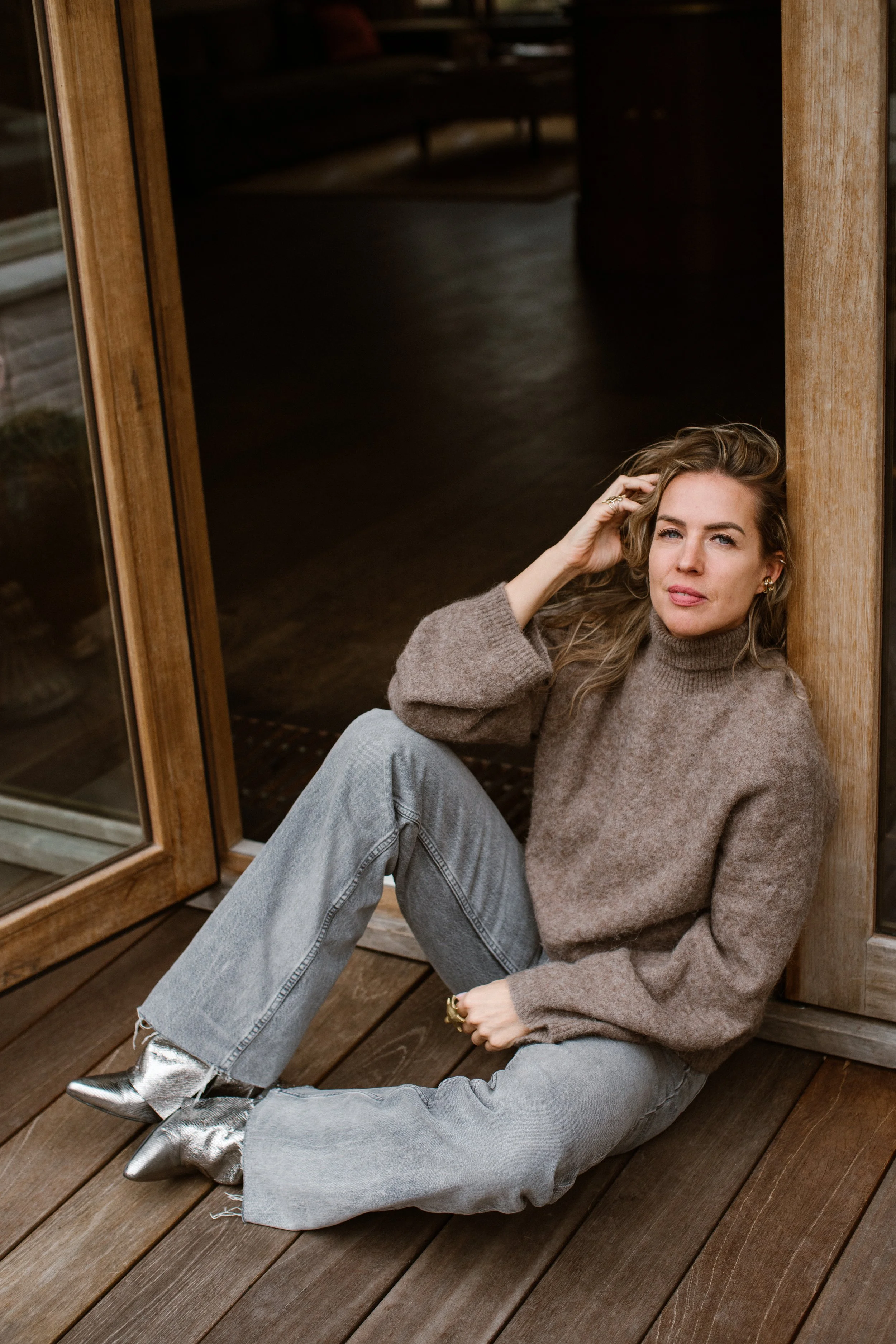 A young woman with wavy hair and light skin sitting on a wooden porch, leaning against a wooden doorframe, wearing a brown sweater, gray jeans, metallic silver ankle boots, and gold jewelry.