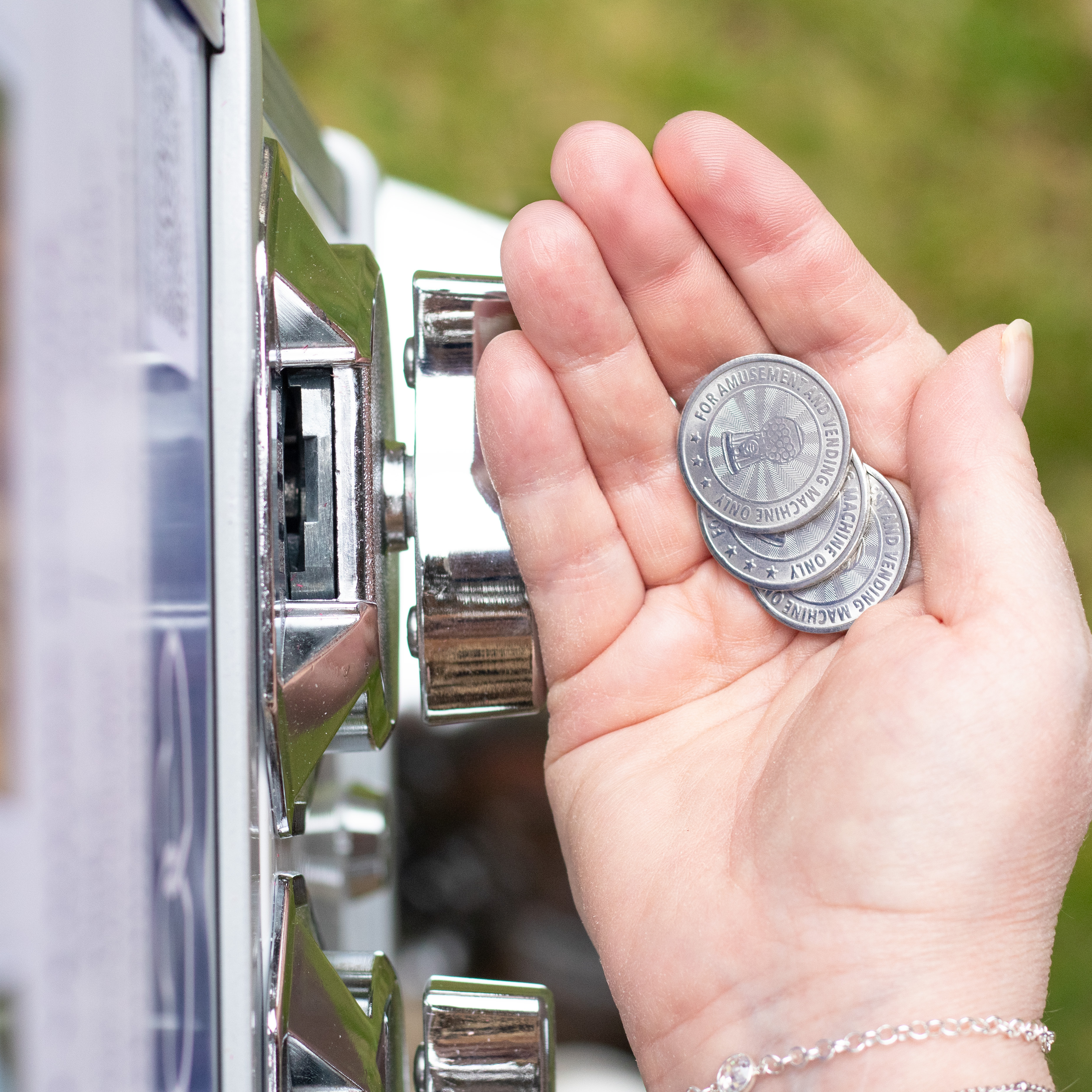 A person's hand with a silver bracelet is inserting a coin into a slot on a metallic vending machine or payment kiosk outdoors, with a blurred green background.