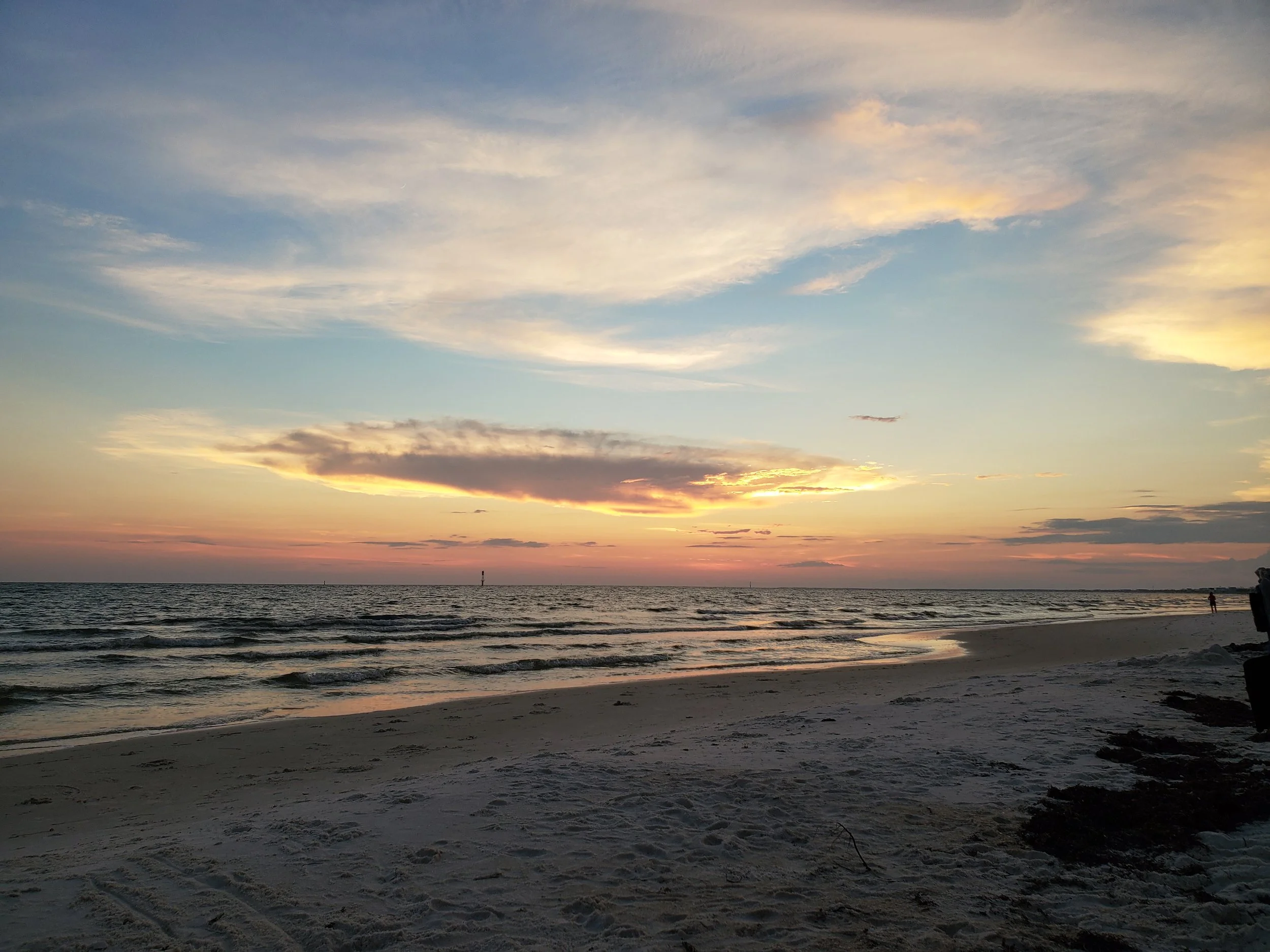 Beach at sunset with a colorful sky, calm ocean waves, and some people walking in the distance.